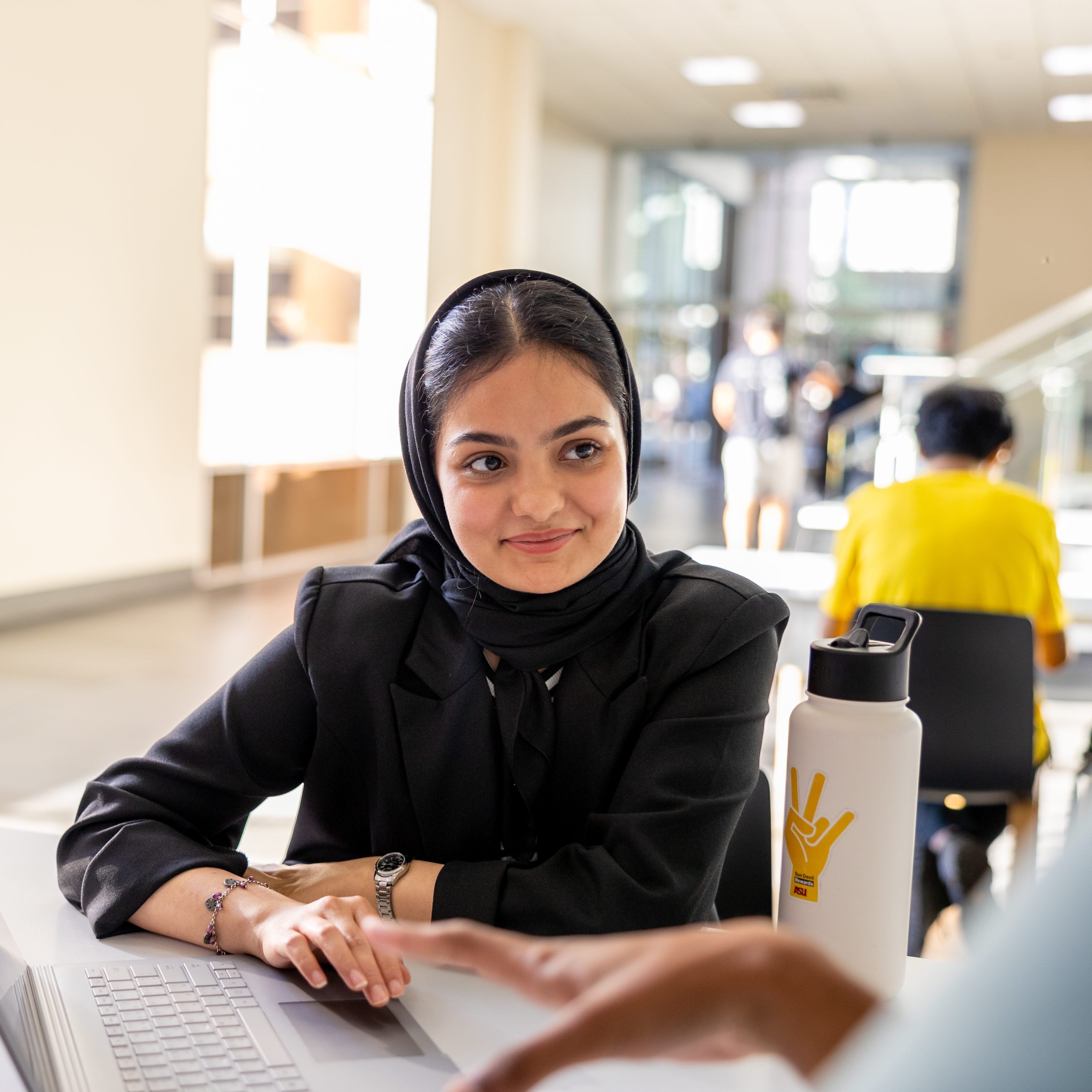 Young woman in a black headscarf sitting at a table with an open laptop and a white water bottle. Another person’s hand is visible in the foreground.