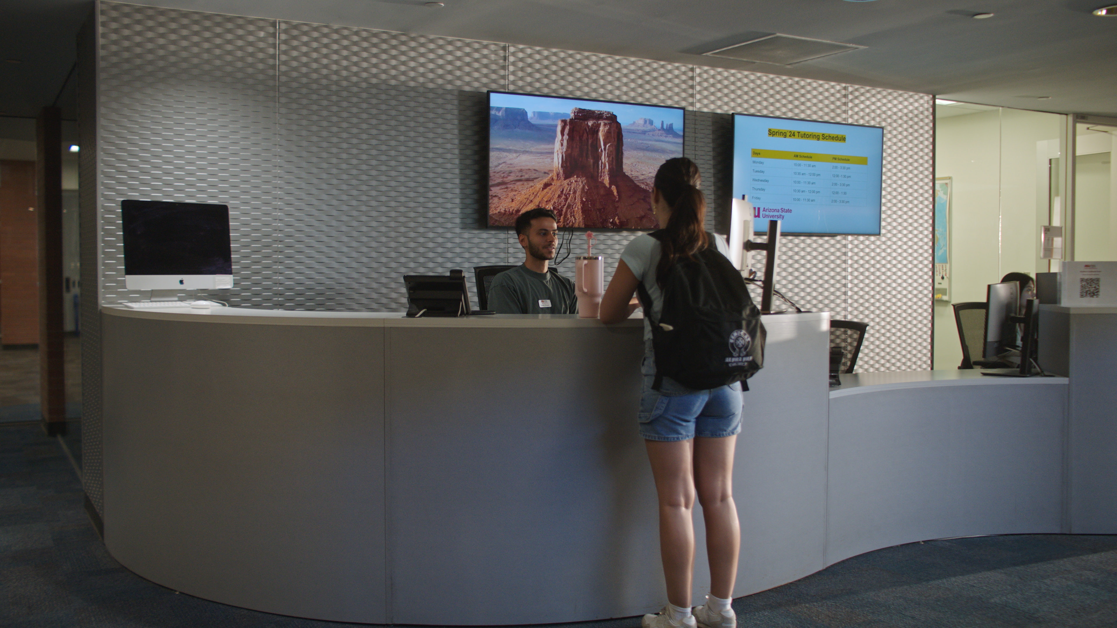 Reception area with a curved grey desk, two people interacting, and two monitors displaying landscape and schedule.