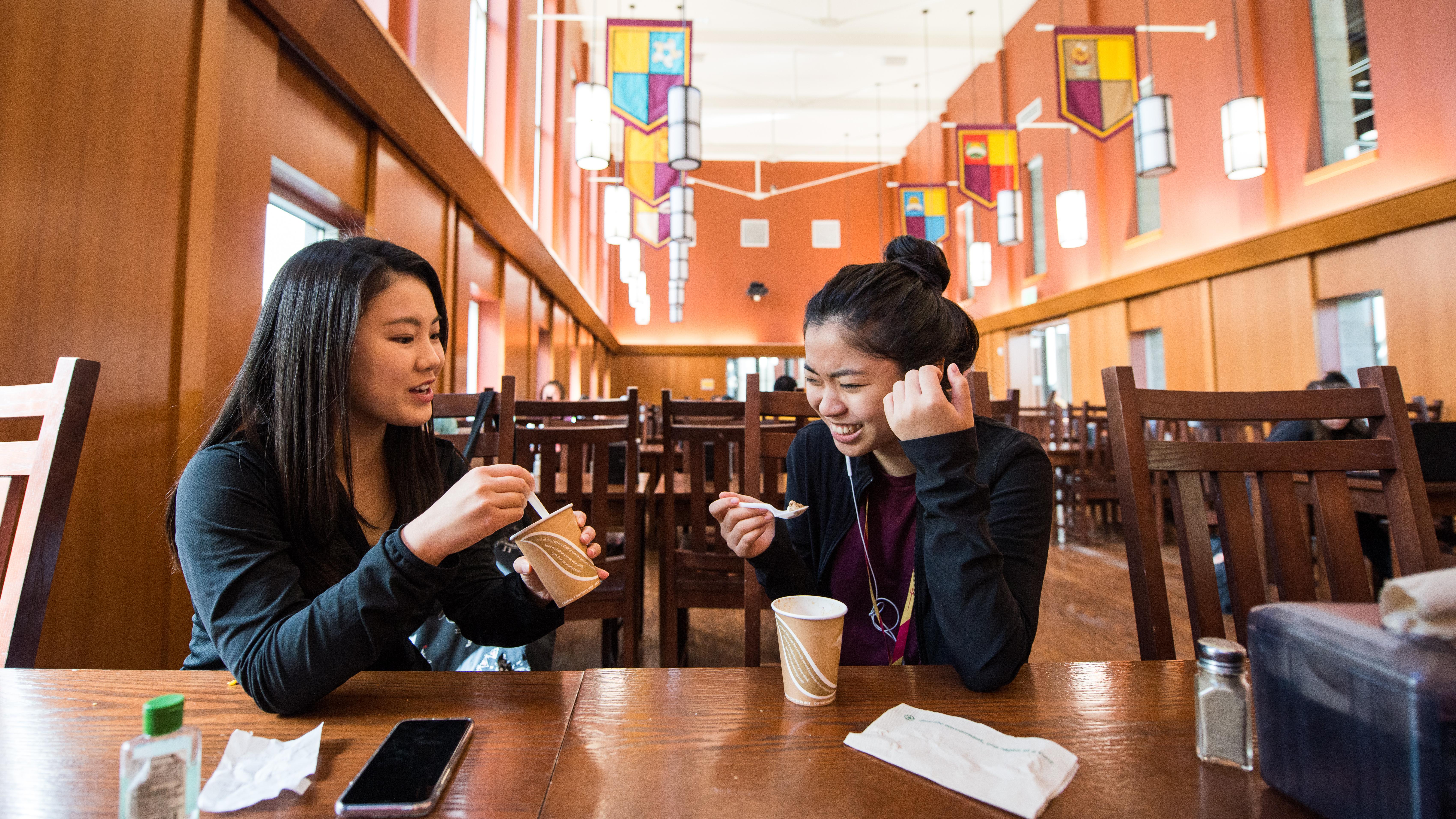 Two students seated at a dining hall table eating and talking.