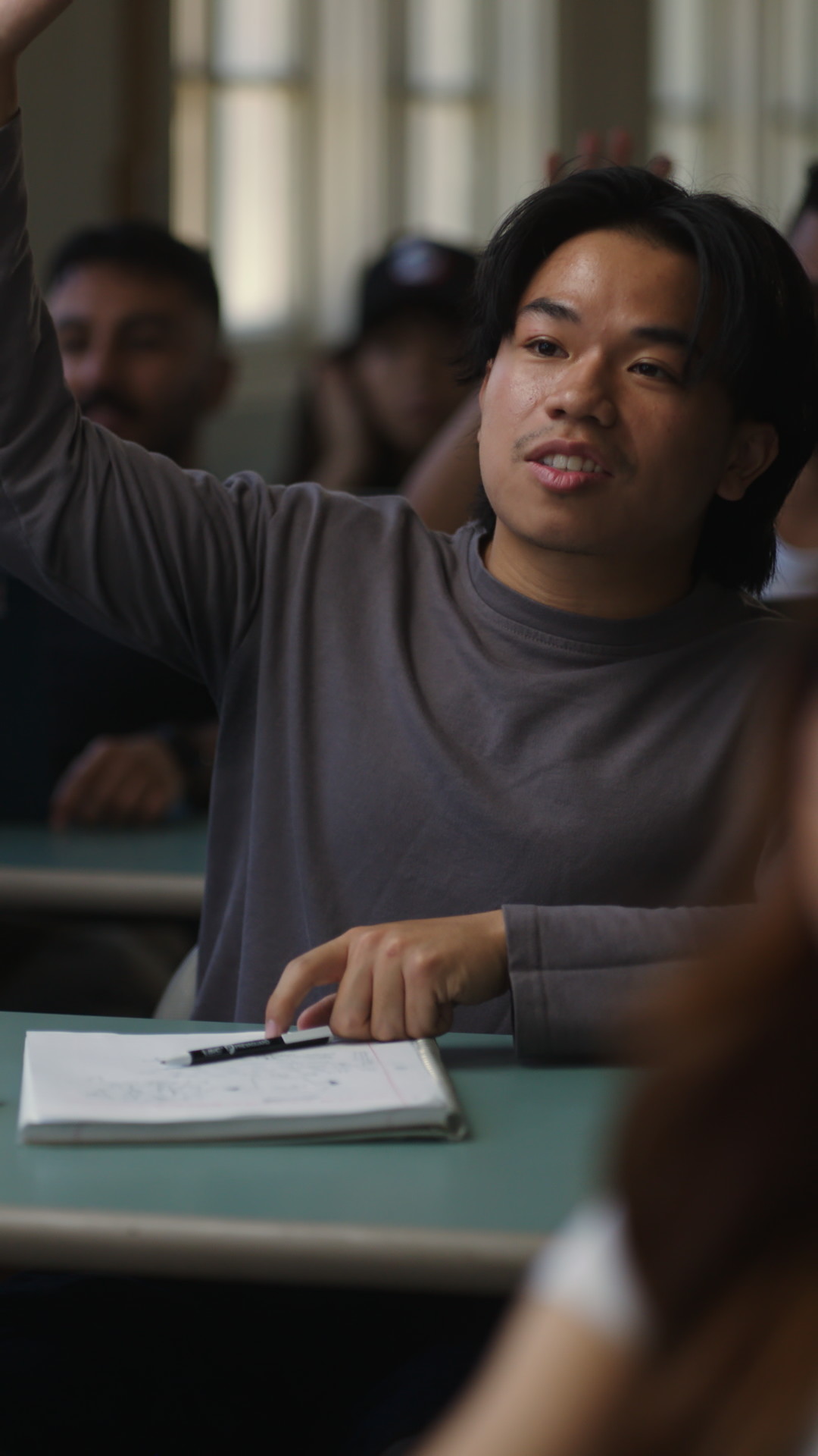 Student in a gray shirt raising their hand in a classroom setting, with a notebook and pen on the desk.