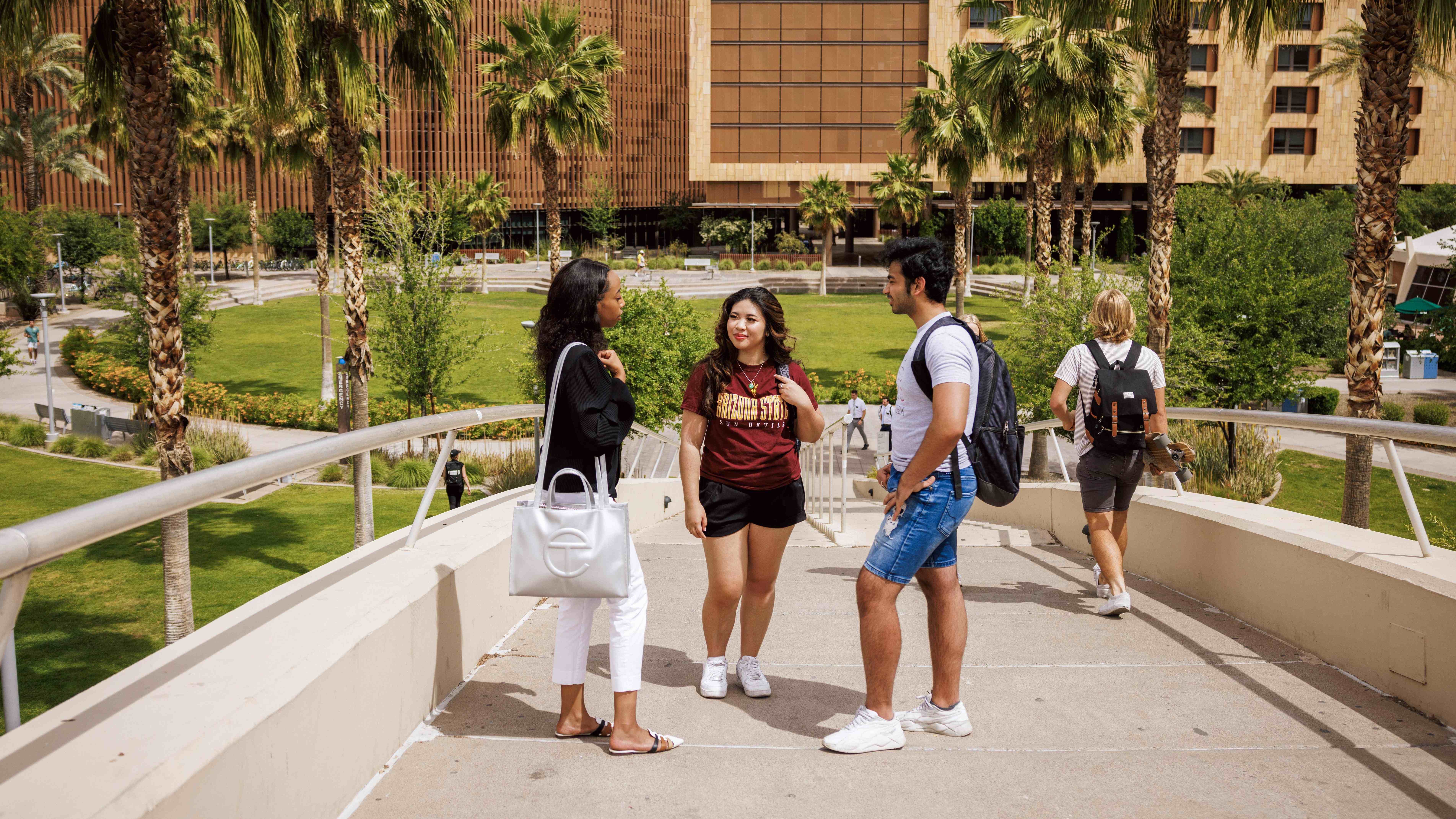 People talking on a campus walkway with palm trees and a large brick building in the background.