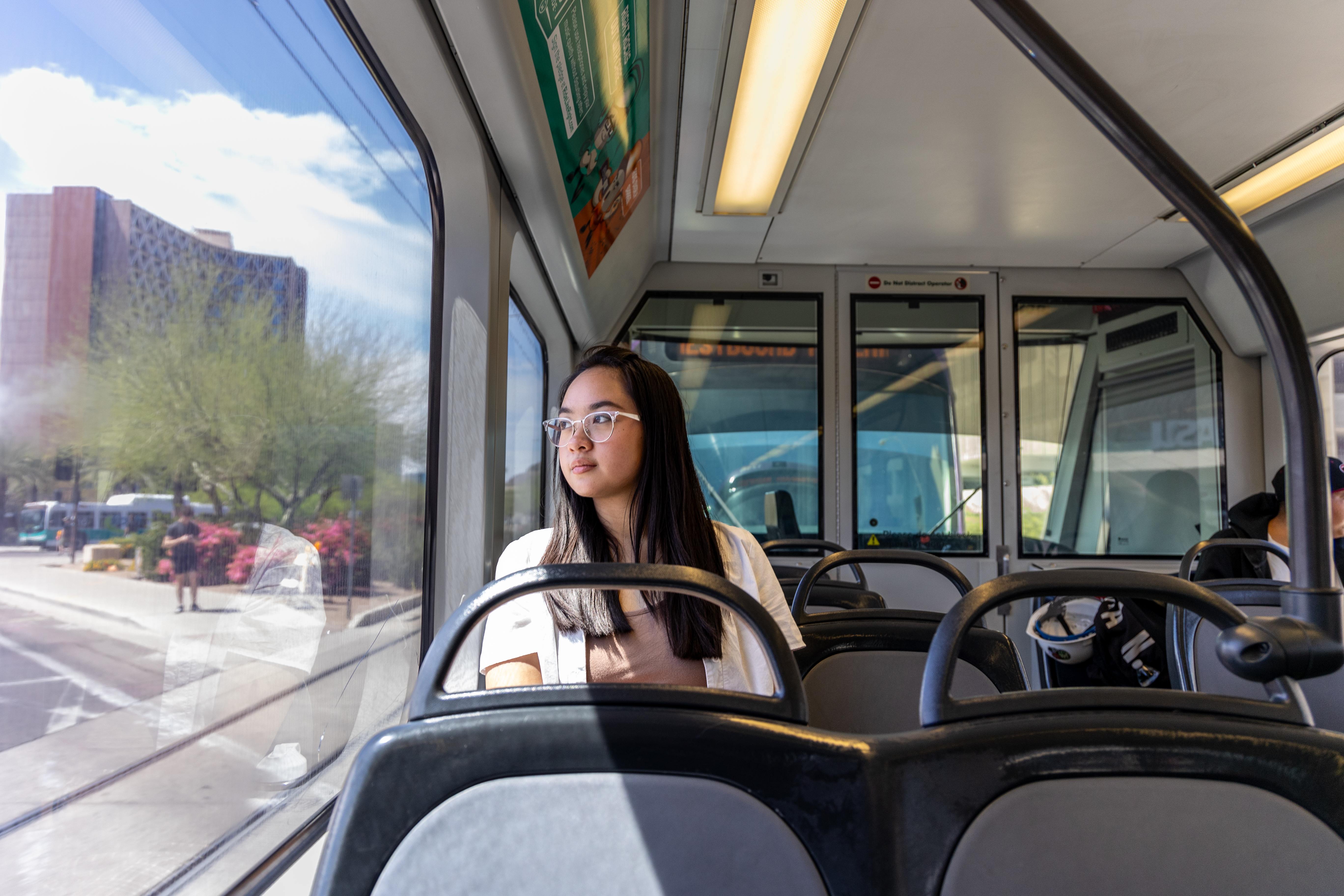 A young student with glasses sits by a window on public transport, gazing outside.