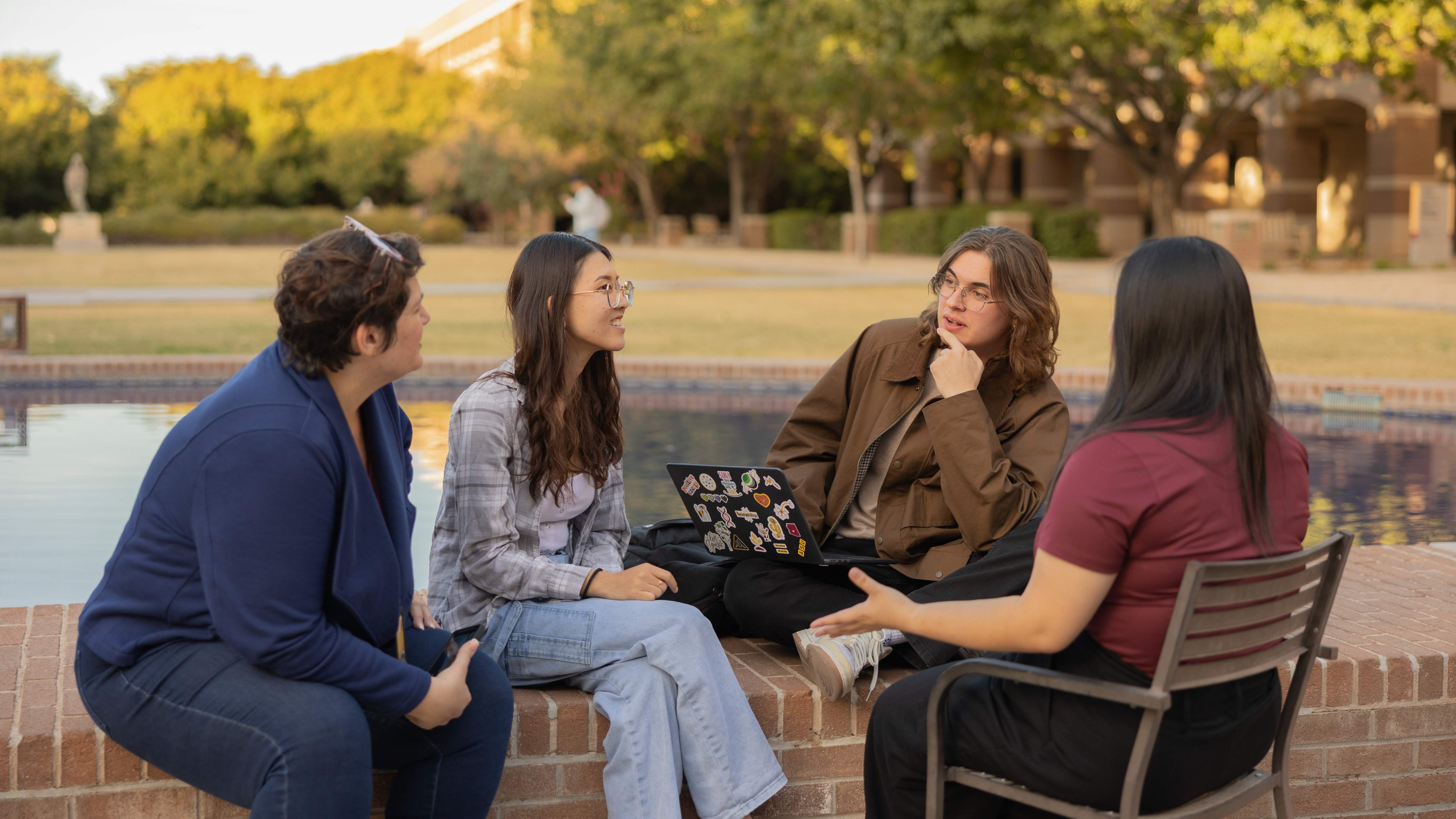 Four people sitting and talking by a brick-lined pool in an outdoor setting.