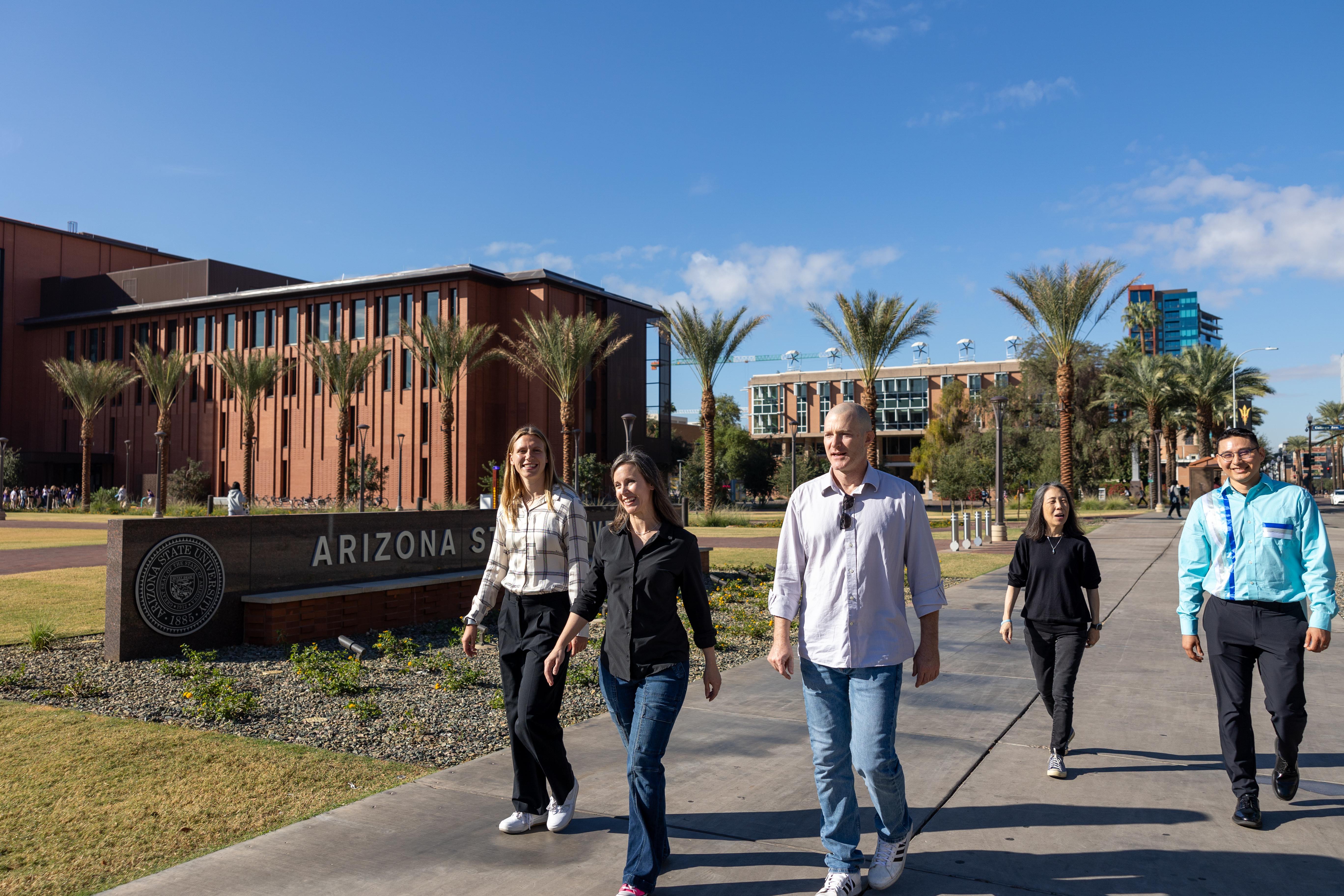 Five people walking on a pathway at Arizona State University with campus buildings and palm trees in the background.