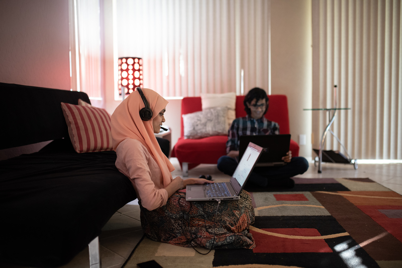 Two students sit on the living room floor of their home, working on their laptops.