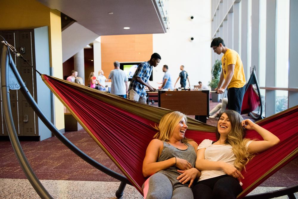 Two laughing ASU students relaxing in a hammock in a game room.​​