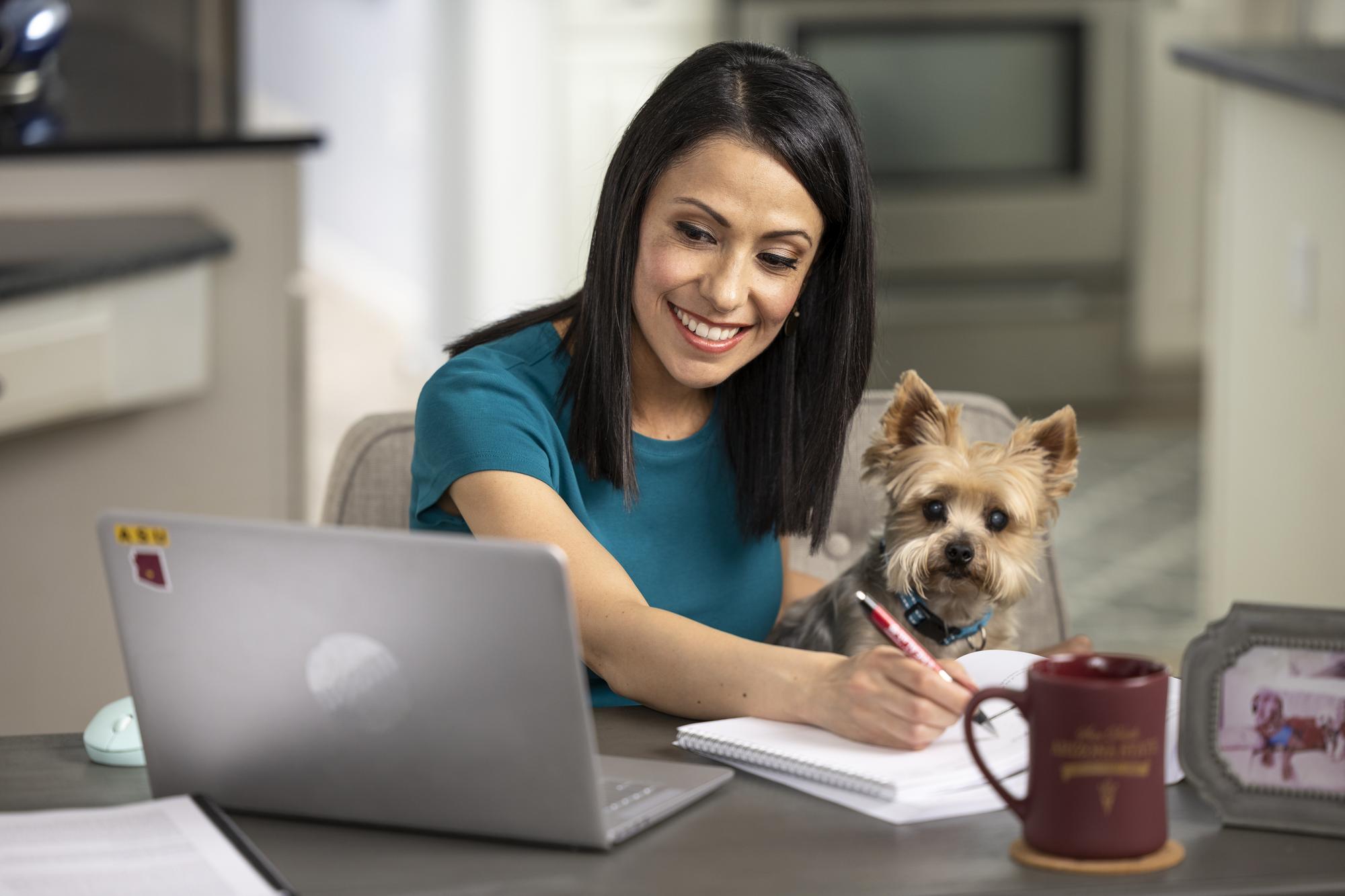 Smiling female ASU student sitting at her laptop with her small dog.