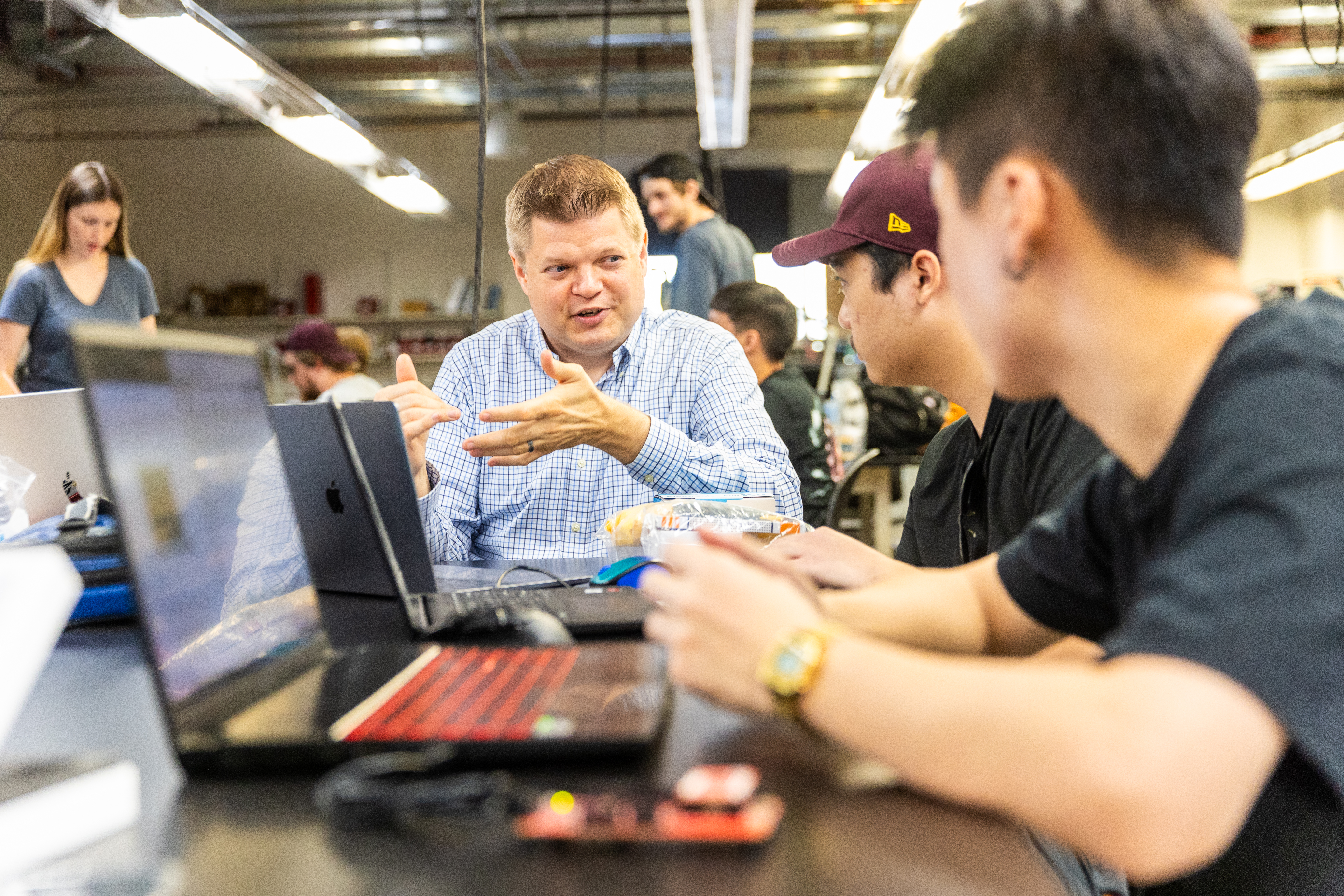 A male instructor explaining directions to two male students sitting at their laptops in class.