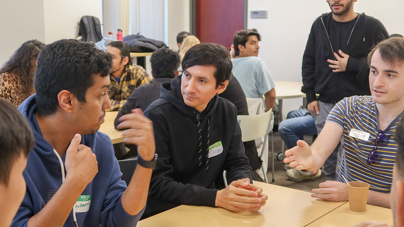 Several students speak and listen during conversation sitting at a desk.