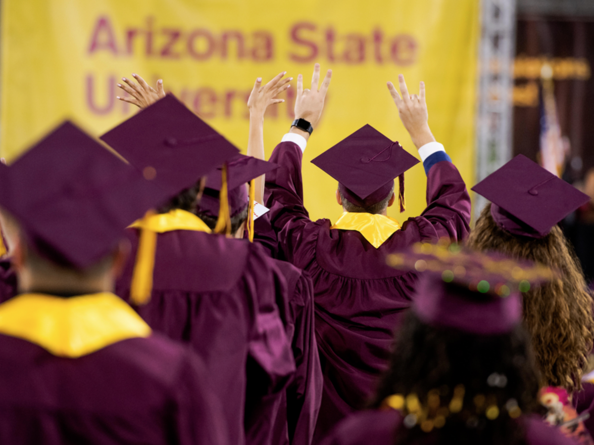 Graduates in maroon caps and gowns raise their hands at a ceremony.