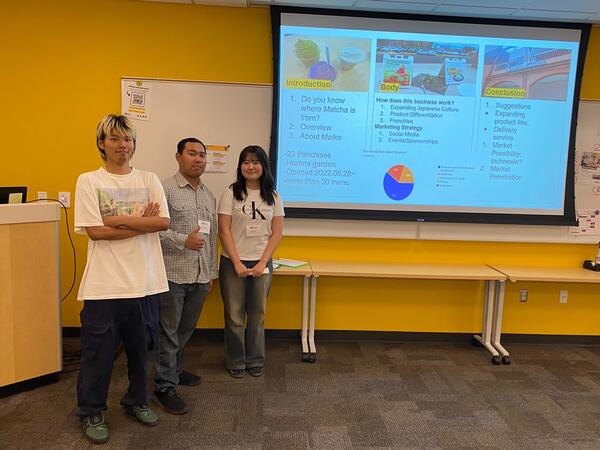 Three people stand in front of a presentation screen in a yellow-walled room.