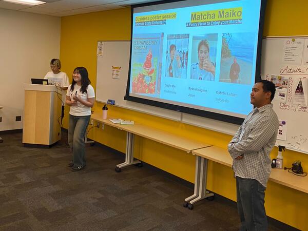 Three people present during a business poster session at Arizona State University. A projection screen displays the topic "Matcha Maiko" and participants' names and countries. A whiteboard to the right has assorted materials pinned.