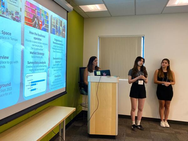 Three young women presenting a slideshow about a craft space in a classroom with a green accent wall.