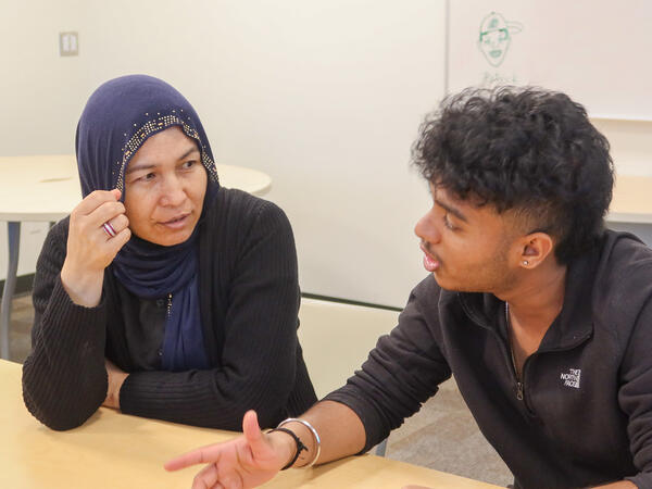 A female student looks at and listens intently to a male student speaking and using his hands to emphasize the topic he is discussing.