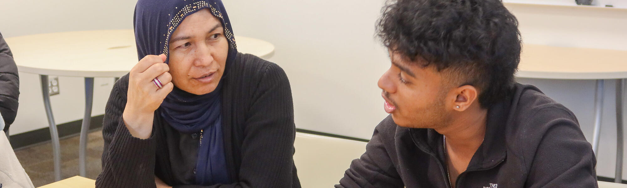 A female student looks at and listens intently to a male student speaking and using his hands to emphasize the topic he is discussing.