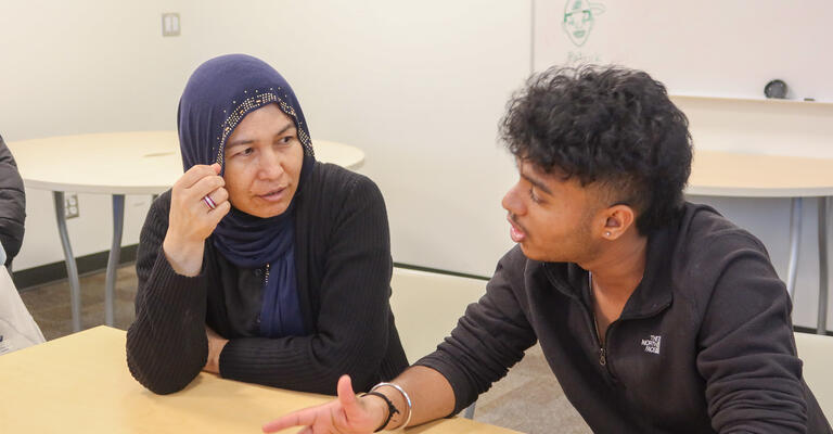 A female student looks at and listens intently to a male student speaking and using his hands to emphasize the topic he is discussing.