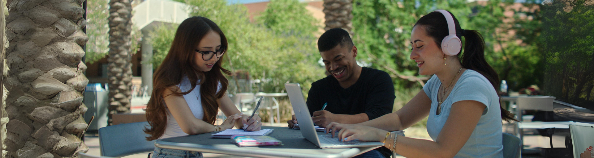 Three people studying at an outdoor table with laptops and notebooks.