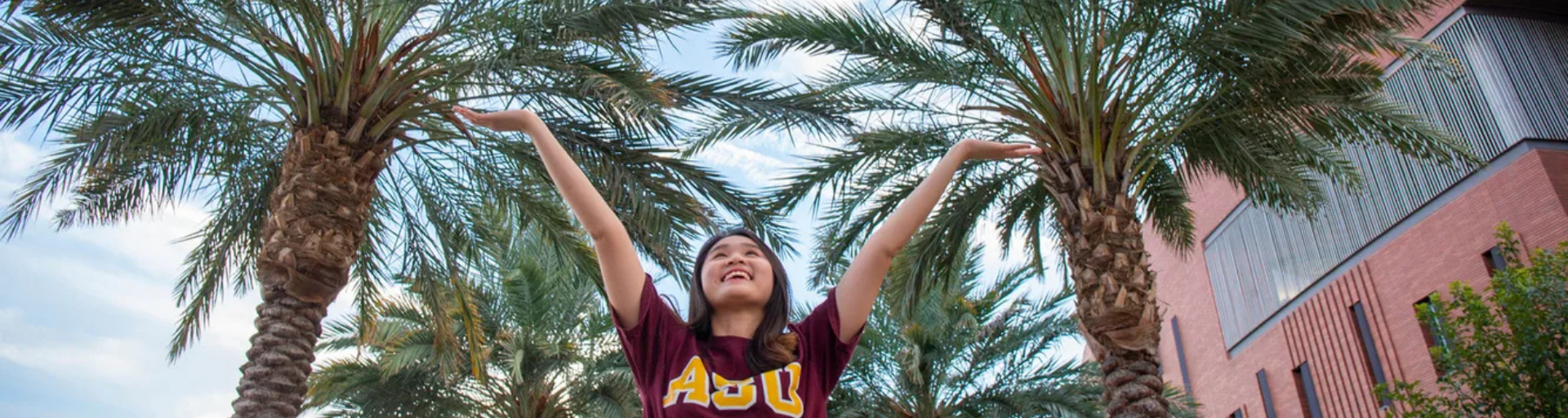 A person in an ASU shirt stands with arms raised between two palm trees, with a building in the background.