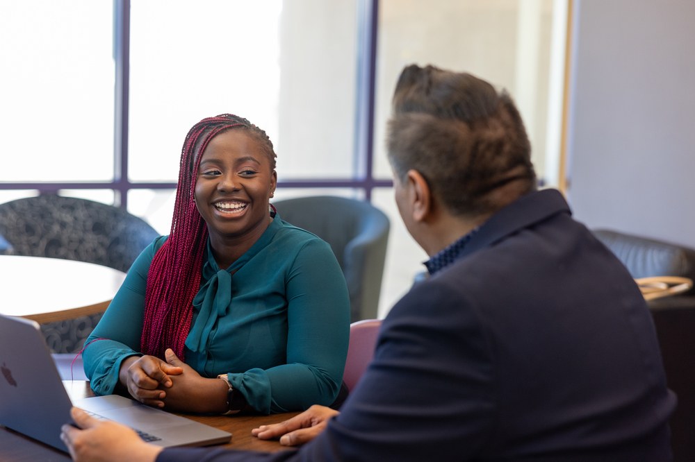 A smiling woman sitting and having a conversation with a man at a desk.